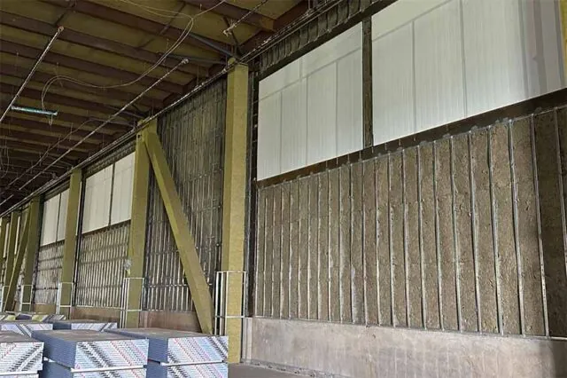Long interior wall retrofit with mineral-wool batts and translucent upper panels; drywall stock on floor.