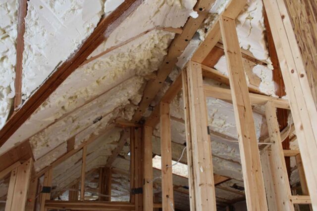 An unfinished attic space under construction, showing off-white spray foam insulation applied between wooden roof rafters and wall studs.